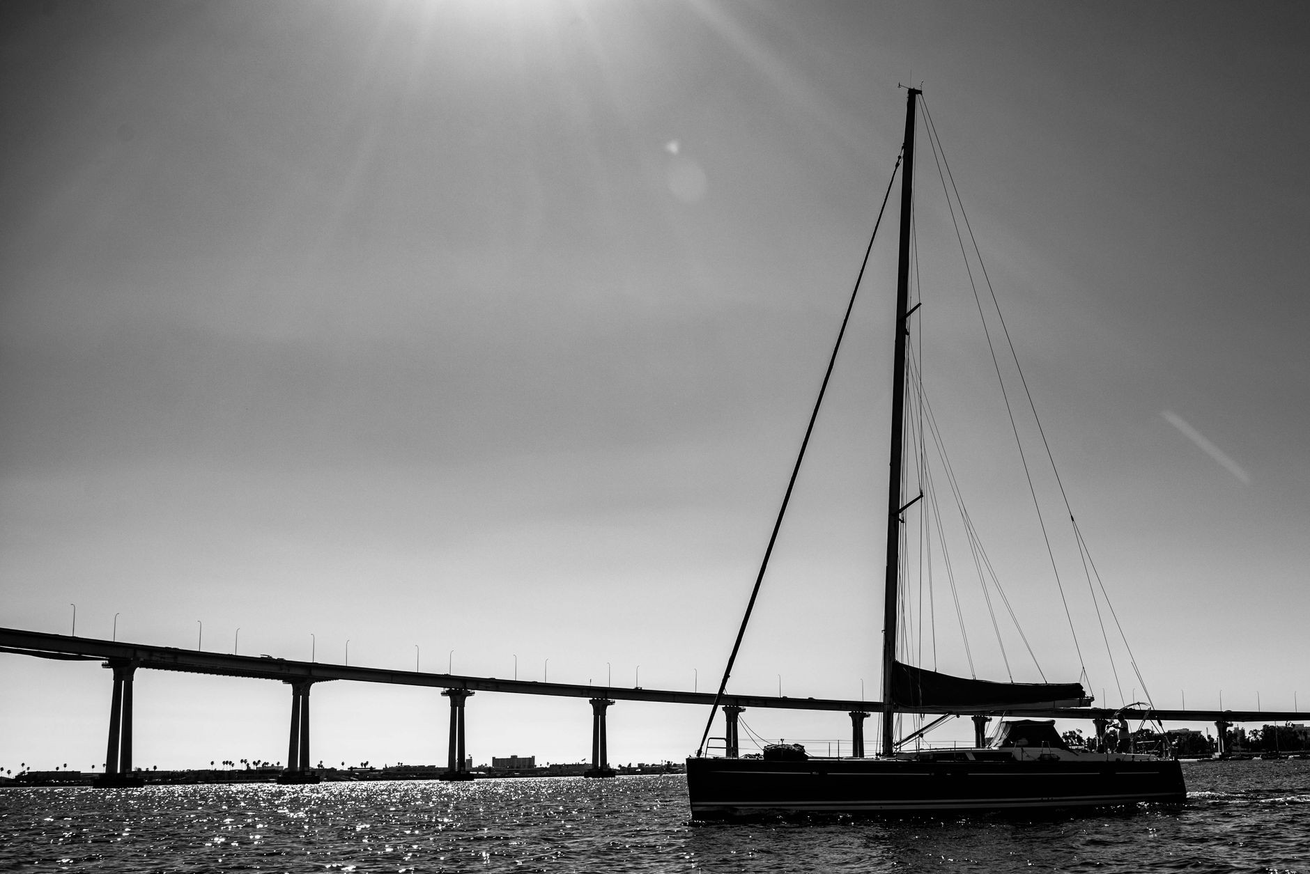 A dramatic black and white photo of a sailboat near a city bridge with sunlight overhead.